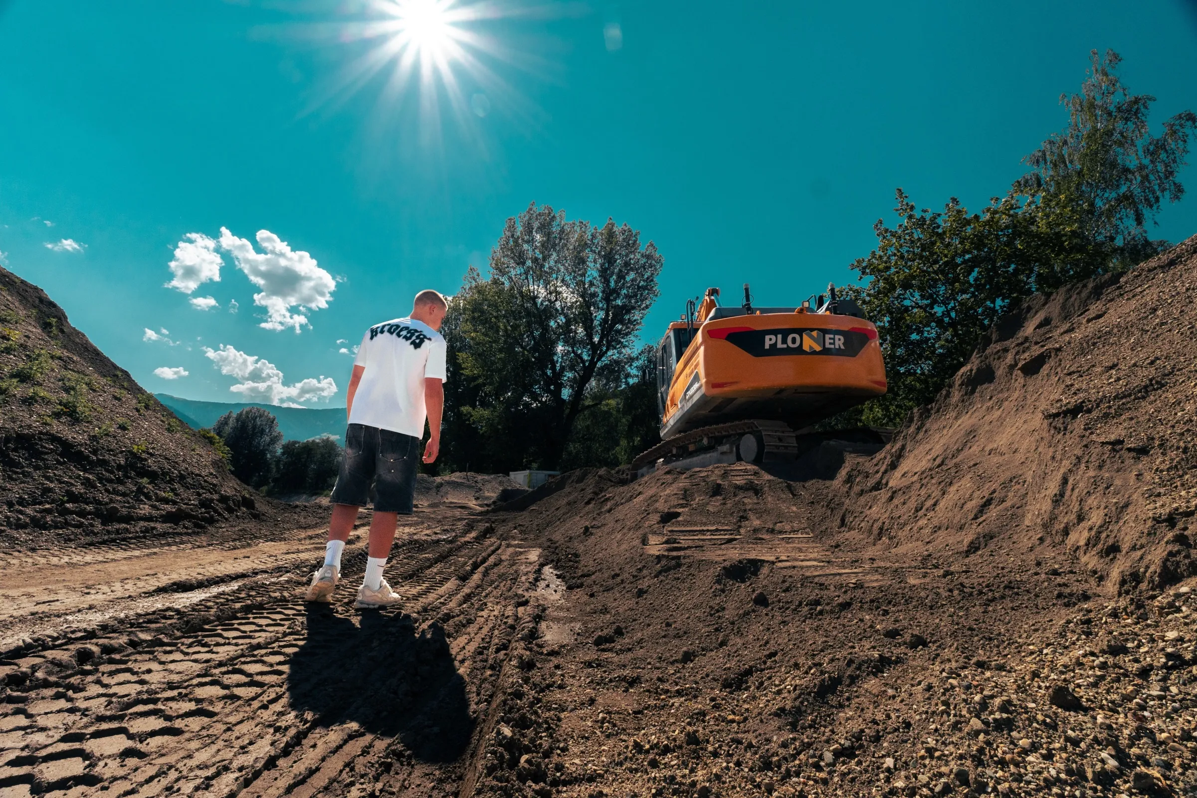 Person in weißem Shirt in einer Sandgrube mit Bagger, Weitwinkelaufnahme gegen die Sonne